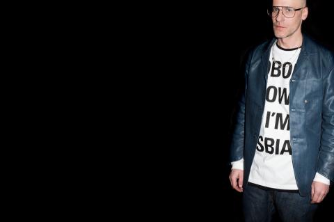 A man with glasses and a printed tshirt is looking into the camera in front of a black background